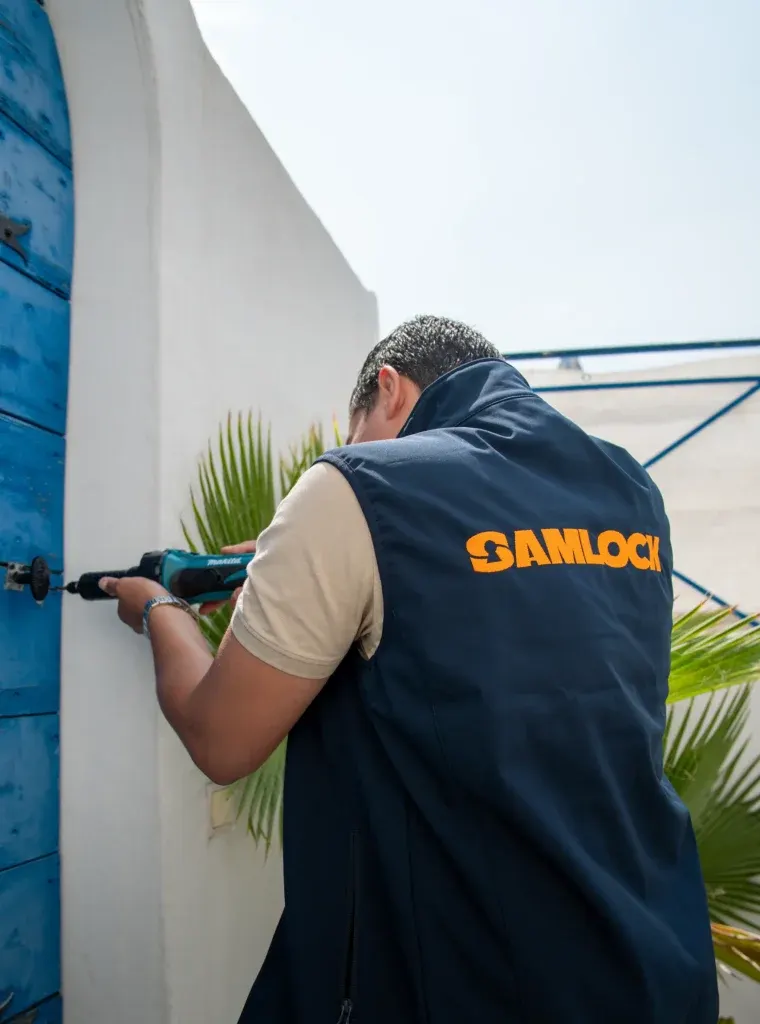 Sami intervient sur la serrure d’une porte bleue avec une visseuse, gilet SamLock sur le dos
